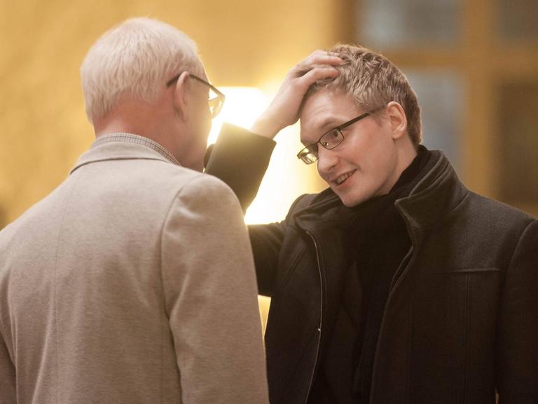 Farbfoto eines jungen blonden Mannes, der sich im Gespräch mit einem älteren Mann befindet, den man nur von hinten sieht