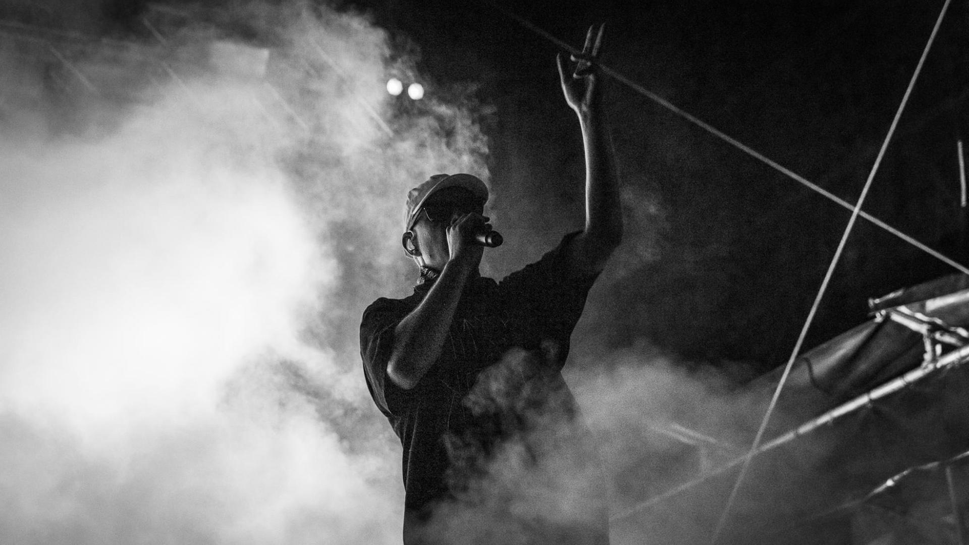 Der Hip-Hop Sänger Trettmann im Bühnen-Nebel, Mikrophon am Mund und Hand in die Luft gestreckt. Der Hip-Hop Sänger Trettmann im Bühnen-Nebel, Mikrophon am Mund und Hand in die Luft gestreckt.