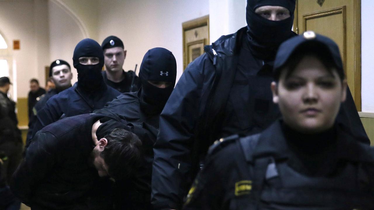 Mehrere Sicherheitskräfte in Uniform und teils vermummt begleiten einen Verdächtigen, der den Kopf gesenkt hält. Mehrere Sicherheitskräfte in Uniform und teils vermummt begleiten einen Verdächtigen, der den Kopf gesenkt hält.
