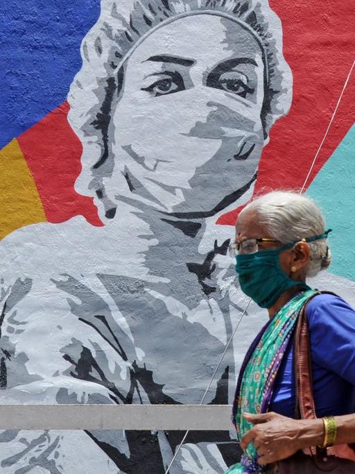 Eine ältere Frau geht an einem Wandgemälde vorbei, auf dem auf farbenfrohem Grund das Schwarzweiß-Bild einer Pflegekraft mit Schutzmaske zu sehen ist. Die Fußgängerin trägt ebenfalls Maske.