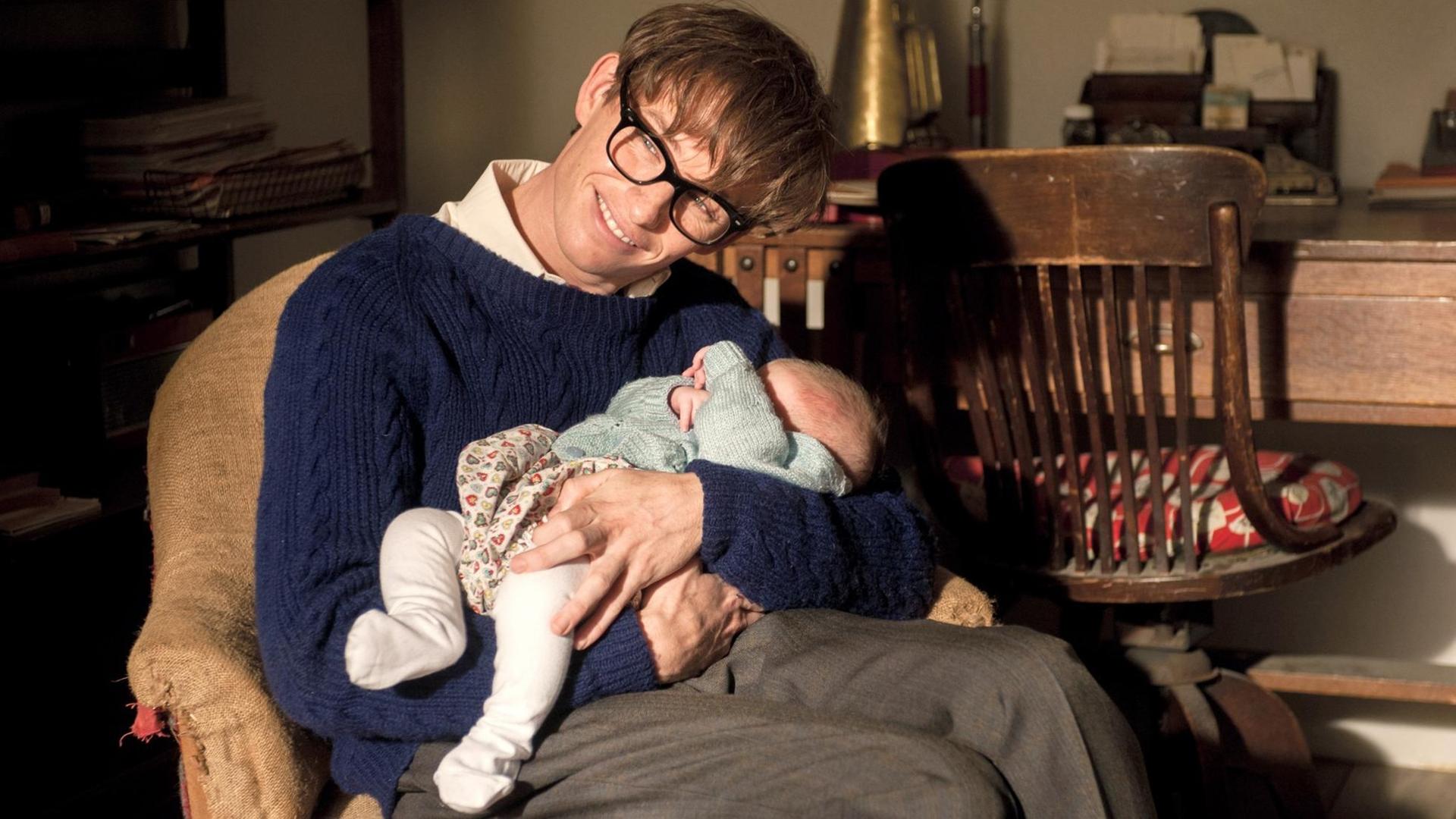 Eddie Redmayne als Stephen Hawking in einer Szene des Kinofilms "Die Entdeckung der Unendlichkeit" (2014). Das Foto zeigt den Hauptdarsteller als schwerbehinderten Physiker Stephen Hawking mit einen Baby auf dem Arm. Eddie Redmayne als Stephen Hawking in einer Szene des Kinofilms "Die Entdeckung der Unendlichkeit" (2014). Das Foto zeigt den Hauptdarsteller als schwerbehinderten Physiker Stephen Hawking mit einen Baby auf dem Arm.