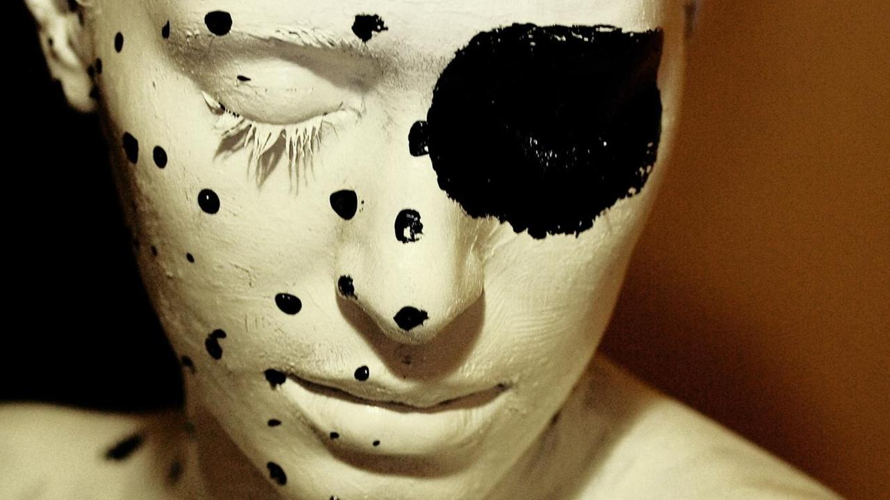 Ein weiß angemaltes Gesicht und Oberkörper, die linke Seite ist mit schwarzes Punkten verziert, über das geschlossene rechte Auge ist ein besonders großer schwarzer Punkt gemalt.