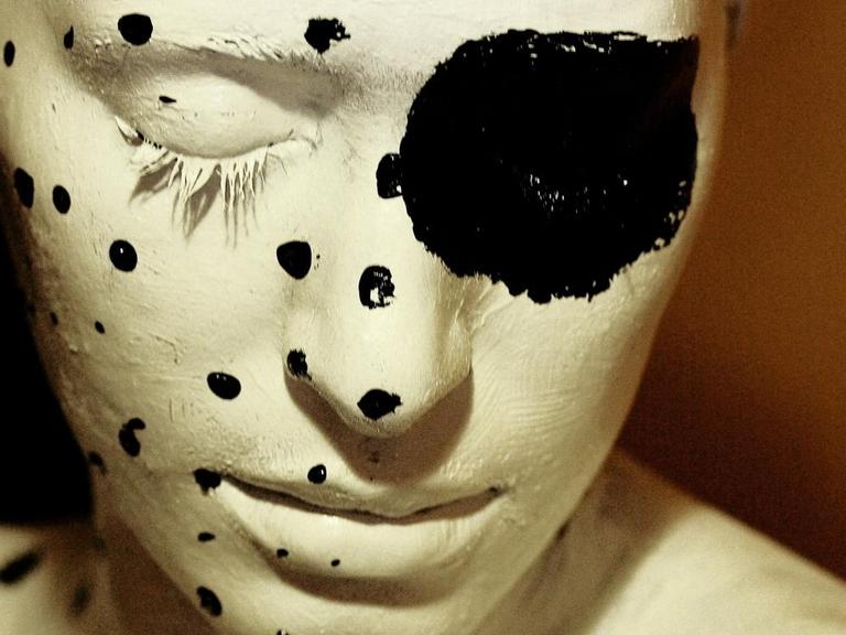Ein weiß angemaltes Gesicht und Oberkörper, die linke Seite ist mit schwarzes Punkten verziert, über das geschlossene rechte Auge ist ein besonders großer schwarzer Punkt gemalt.