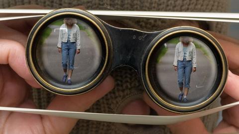 Das gestellte Foto zeigt eine maskierte Person, die mit einem Fernglas durch die Lamellen einer Jalousie hindurch eine Frau beobachtet.