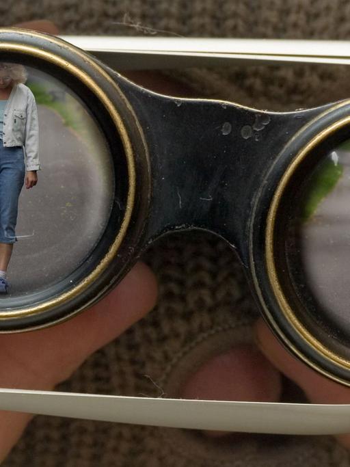 Das gestellte Foto zeigt eine maskierte Person, die mit einem Fernglas durch die Lamellen einer Jalousie hindurch eine Frau beobachtet