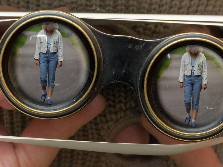 Das gestellte Foto zeigt eine maskierte Person, die mit einem Fernglas durch die Lamellen einer Jalousie hindurch eine Frau beobachtet.