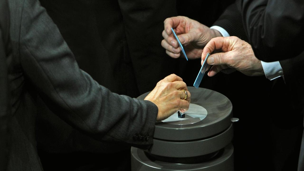 Drei Hände stecken Stimmkarten in eine Wahlurne - namentliche Abstimmung im Bundestag