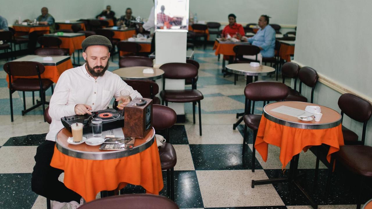 Ein Mann mit Schiebermütze, beigem Leinenhemd und Bart sitzt an einem kleinen runden Tisch, vor sich ein Kaffee. Mit der rechten Hand hebt er den Tonarm eines tragbaren Plattenspielers auf eine schwarze Vinyl-Single.