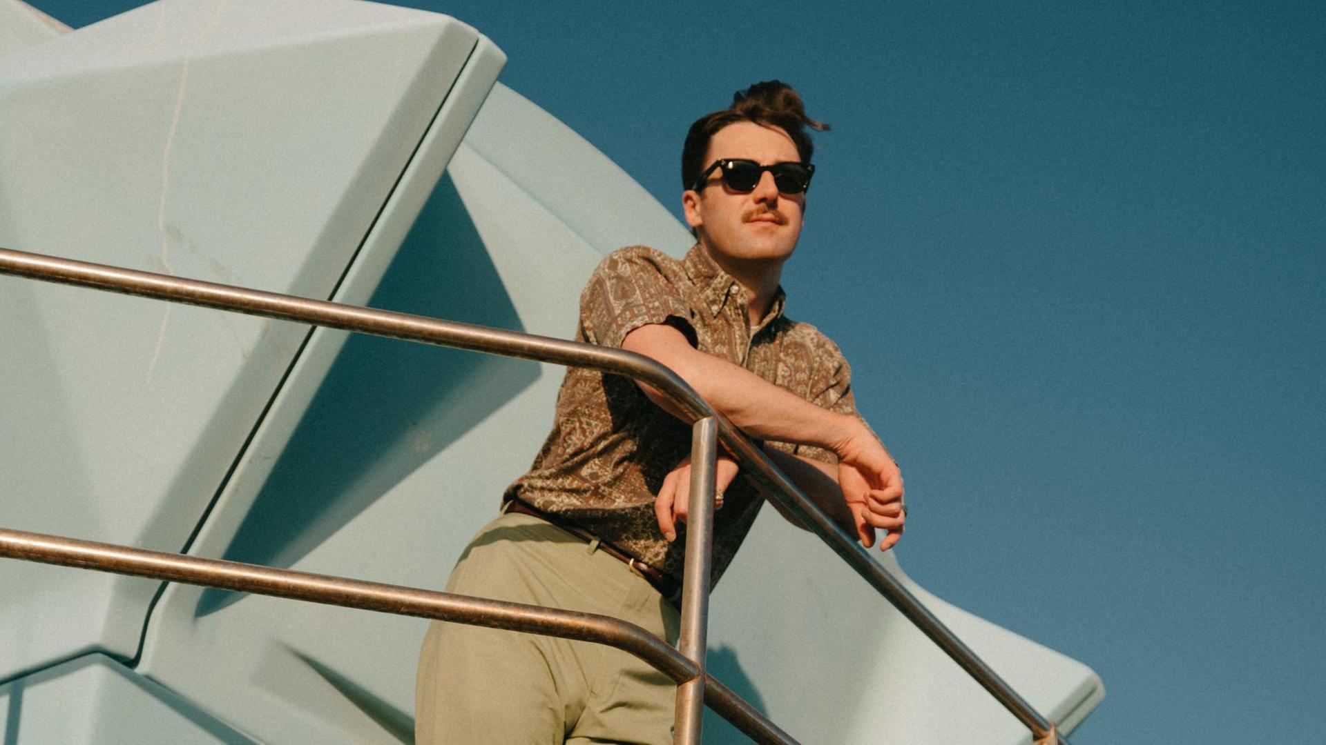Ein Mann mit Sonnenbrille lehnt mit den Armen auf einem Geländer und blickt in die Ferne. Ein Mann mit Sonnenbrille lehnt mit den Armen auf einem Geländer und blickt in die Ferne.