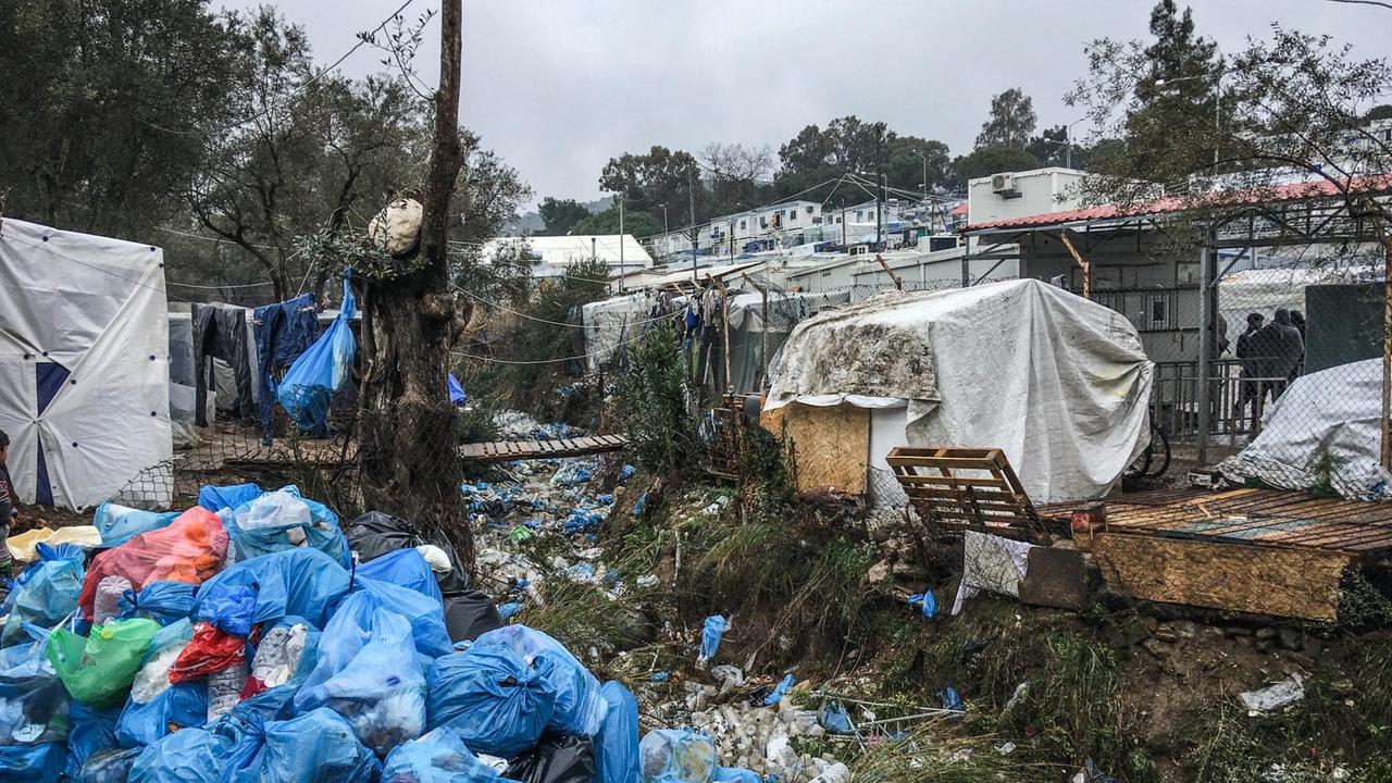 Zu sehen ist eine informelle Wohnstruktur aus Zelten, Containern und selbst gebauten Holzhütten. Dazwischen ein Graben, in dem Unmengen an leeren Plastikfalschen liegen, im Vordergrund links stapeln sich blaue Müllsäcke. Zu sehen ist eine informelle Wohnstruktur aus Zelten, Containern und selbst gebauten Holzhütten. Dazwischen ein Graben, in dem Unmengen an leeren Plastikfalschen liegen, im Vordergrund links stapeln sich blaue Müllsäcke.