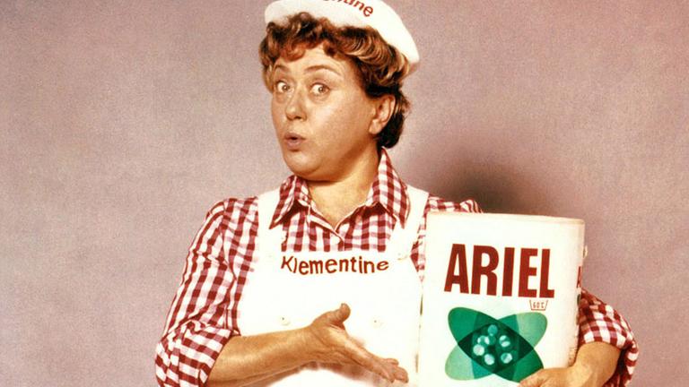 Frau Klementine warb seit 1968 für das Waschmittel Ariel. Sie trägt eine weiße Mütze, einen weißen Overal mit ihrem Namen darauf und hält eine Packung Waschmittel in Händen.