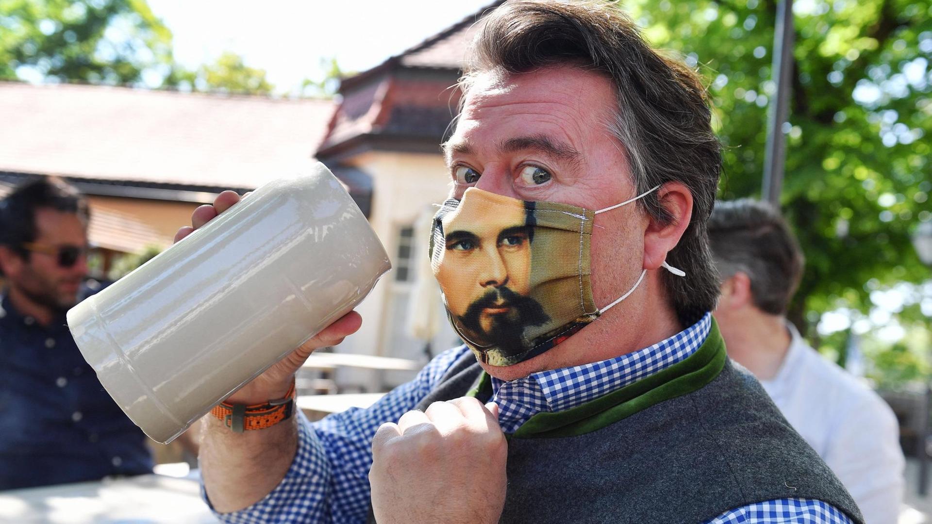 Ein Mann hebt einen Maßkrug, während er eine Gesichtsmaske mit einem Bild von König Ludwig trägt. Ein Mann hebt einen Maßkrug, während er eine Gesichtsmaske mit einem Bild von König Ludwig trägt.