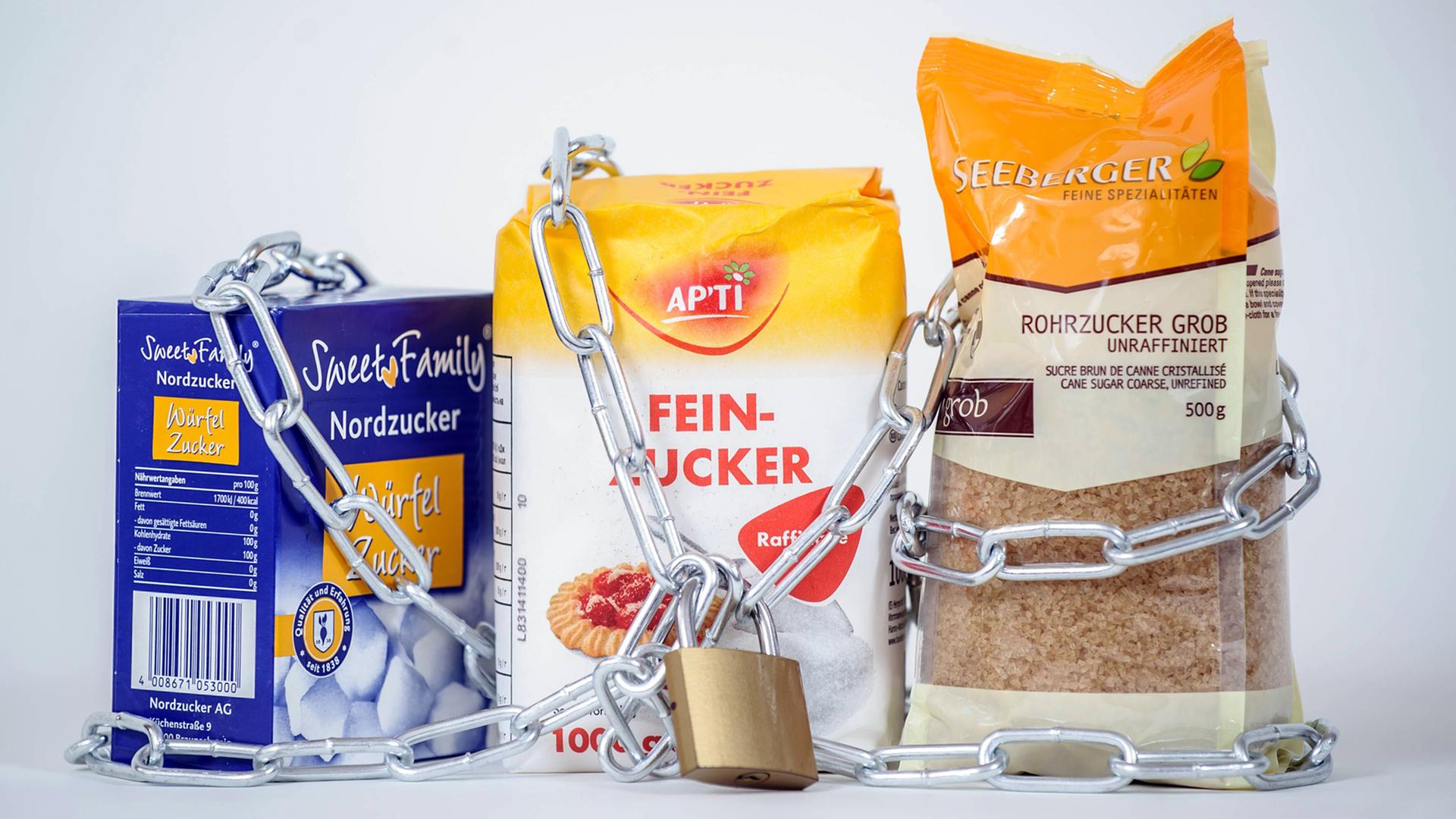 Eine Packung weißer Zucker (M), Würfelzucker (l) und brauner Rohzucker werden von einer Eisenkette samt Schloss zusammen gehalten. Eine Packung weißer Zucker (M), Würfelzucker (l) und brauner Rohzucker werden von einer Eisenkette samt Schloss zusammen gehalten.