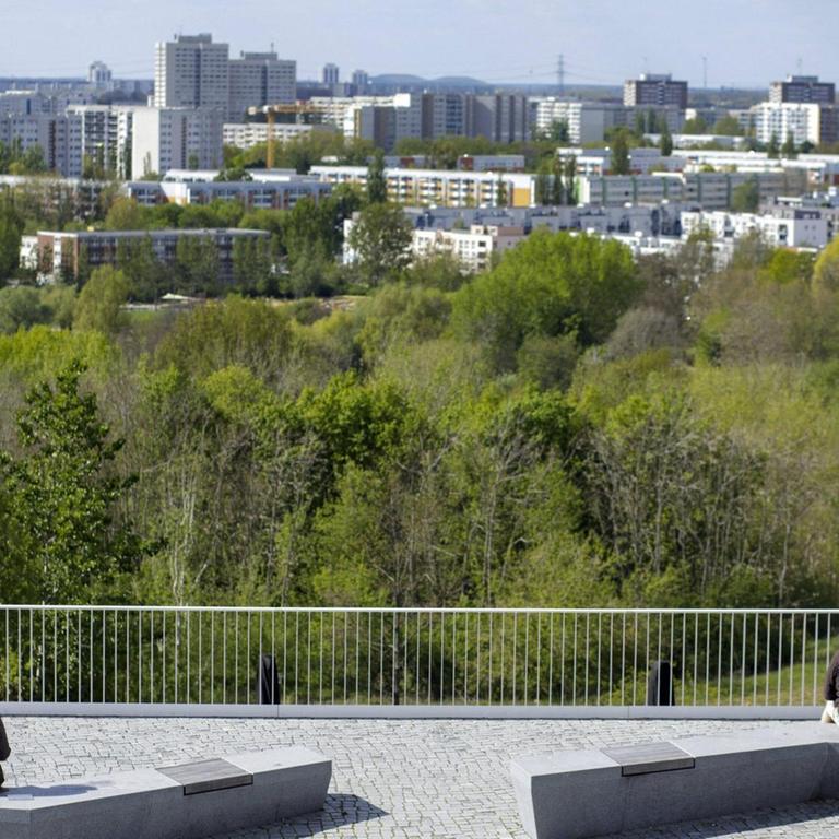 Zwei Paare sitzen am Stadtrand jeweils auf einer Bank mit viel Abstand zueinander und blicken auf die Stadt Berlin in der Ferne. 