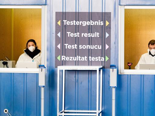 Zwei Personen in Schutzanzügen stehen an der größten Teststrasse Österreichs. Mehrsprachig ist "Testergebnis" zwischen den Fenstern geschrieben.