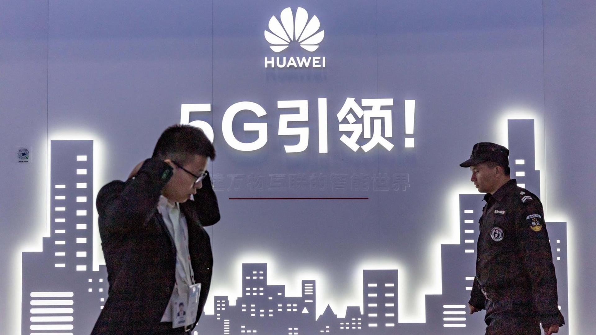 An der Wand eine blaue Grafik mit Hochhäusern, darüber stehen mit weißer Schrift Huawei und 5G sowie chinesische Schriftzeichen. Vor der Wand links ein Mann, der im Laufen telefoniert, von rechts kommt ein Uniformierter. An der Wand eine blaue Grafik mit Hochhäusern, darüber stehen mit weißer Schrift Huawei und 5G sowie chinesische Schriftzeichen. Vor der Wand links ein Mann, der im Laufen telefoniert, von rechts kommt ein Uniformierter.