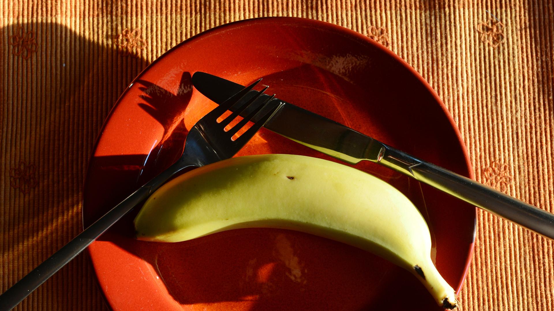 Besteck und eine Banane liegen auf einem orangefarbenen Teller. Besteck und eine Banane liegen auf einem orangefarbenen Teller.