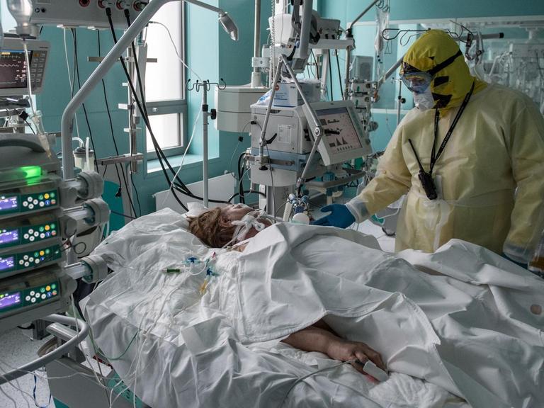 Der Arzt in voller Schutzkleidung steht an der Bettseite der Patientin, die unter einer weißen Decke liegt. Um sie herum Geräte, Kabel und Schläuche.