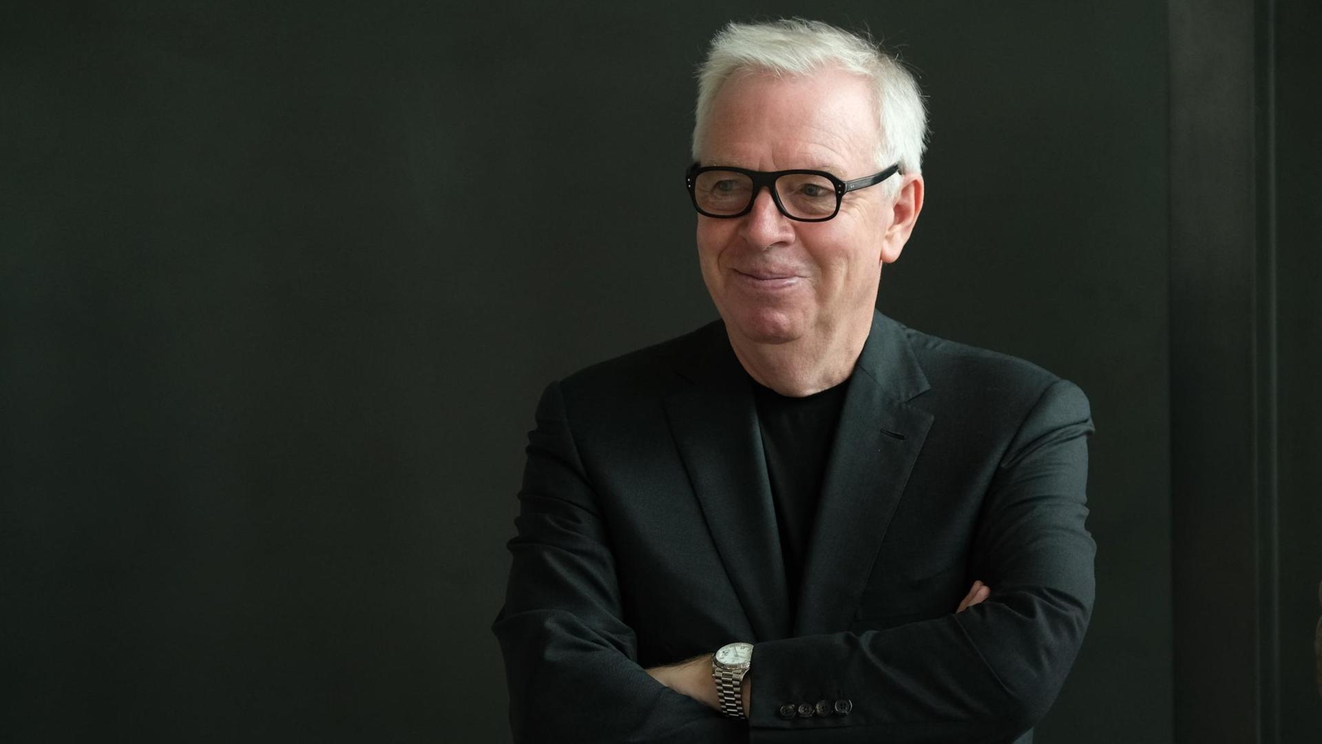 David Chipperfield steht im schwarzen Anzug mit verschränkten Armen vor schwarzem Hintergrund. David Chipperfield steht im schwarzen Anzug mit verschränkten Armen vor schwarzem Hintergrund.