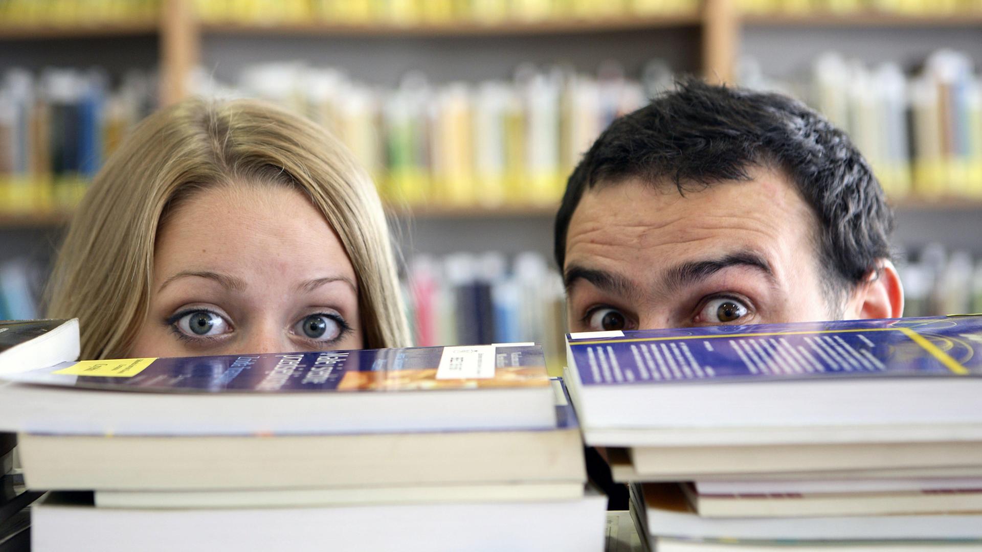 Ein Man und eine Frau verstecken sich hinter einem Stapel Bücher. Von den Gesichtern sind nur die großen offenen Augen und der Haaransatz zu sehen. Ein Man und eine Frau verstecken sich hinter einem Stapel Bücher. Von den Gesichtern sind nur die großen offenen Augen und der Haaransatz zu sehen.