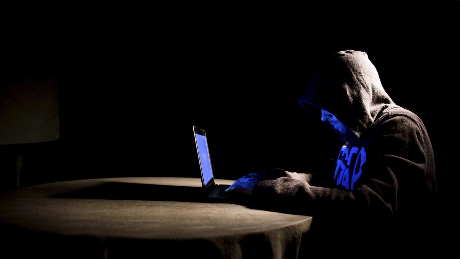 Mann mit Kapuze über dem Kopf sitzt bei sehr schwacher Beleuchtung an einem Tisch. Auf dem Tisch steht ein Laptop. Mann mit Kapuze über dem Kopf sitzt bei sehr schwacher Beleuchtung an einem Tisch. Auf dem Tisch steht ein Laptop.