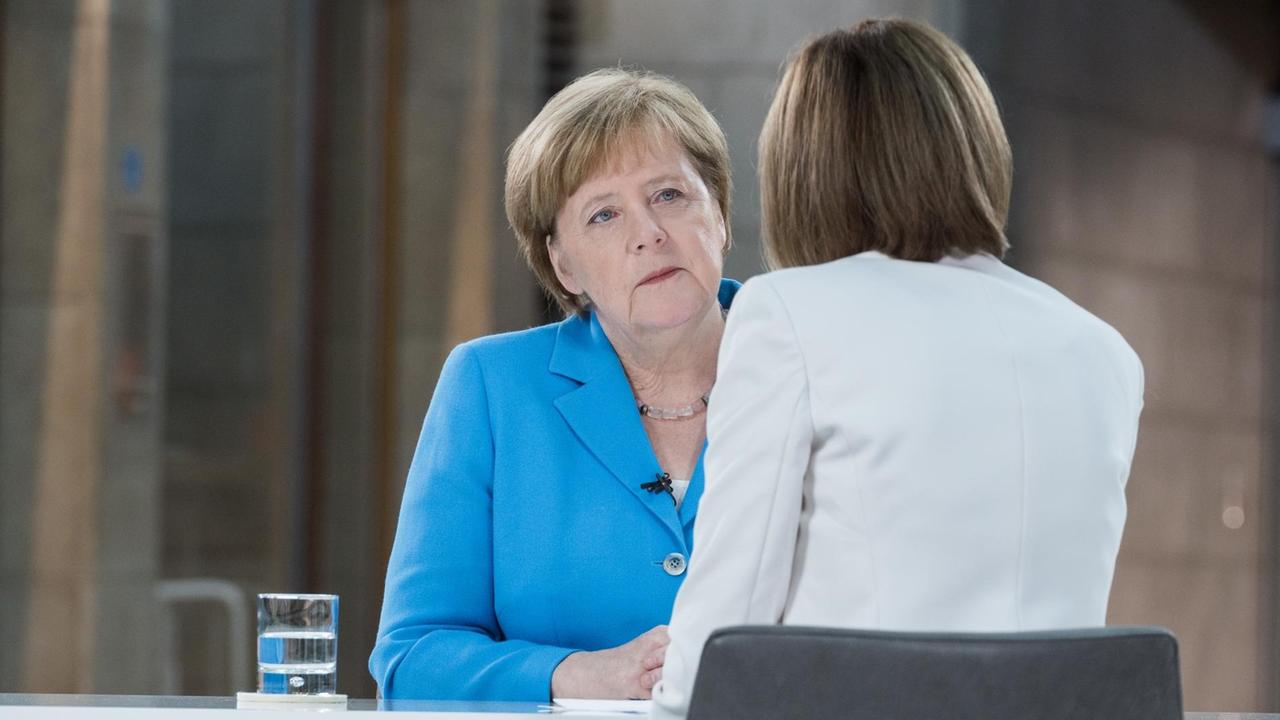 Die Interviewerin im weißen Jackett sieht man von hinten an einem Tisch sitzen. Ihr gegenüber sitzt Merkel im blauen Jackett und hört aufmerksam zu.
