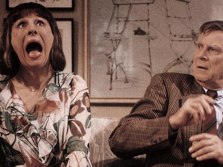 Eine Frau reißt ihren Mund weit auf, der Mann neben ihr auf dem Sofa schreckt zurück.