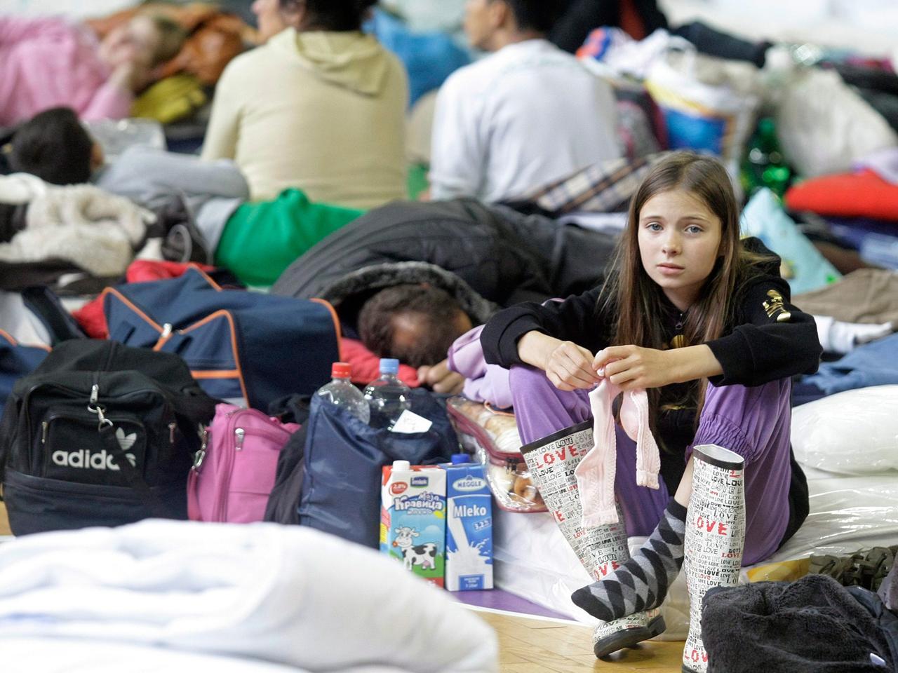 Ein Mädchen sitzt in einer Notunterkunft in Serbien. Sie ist umgeben von Menschen, die auf dem Boden liegen und sitzen.