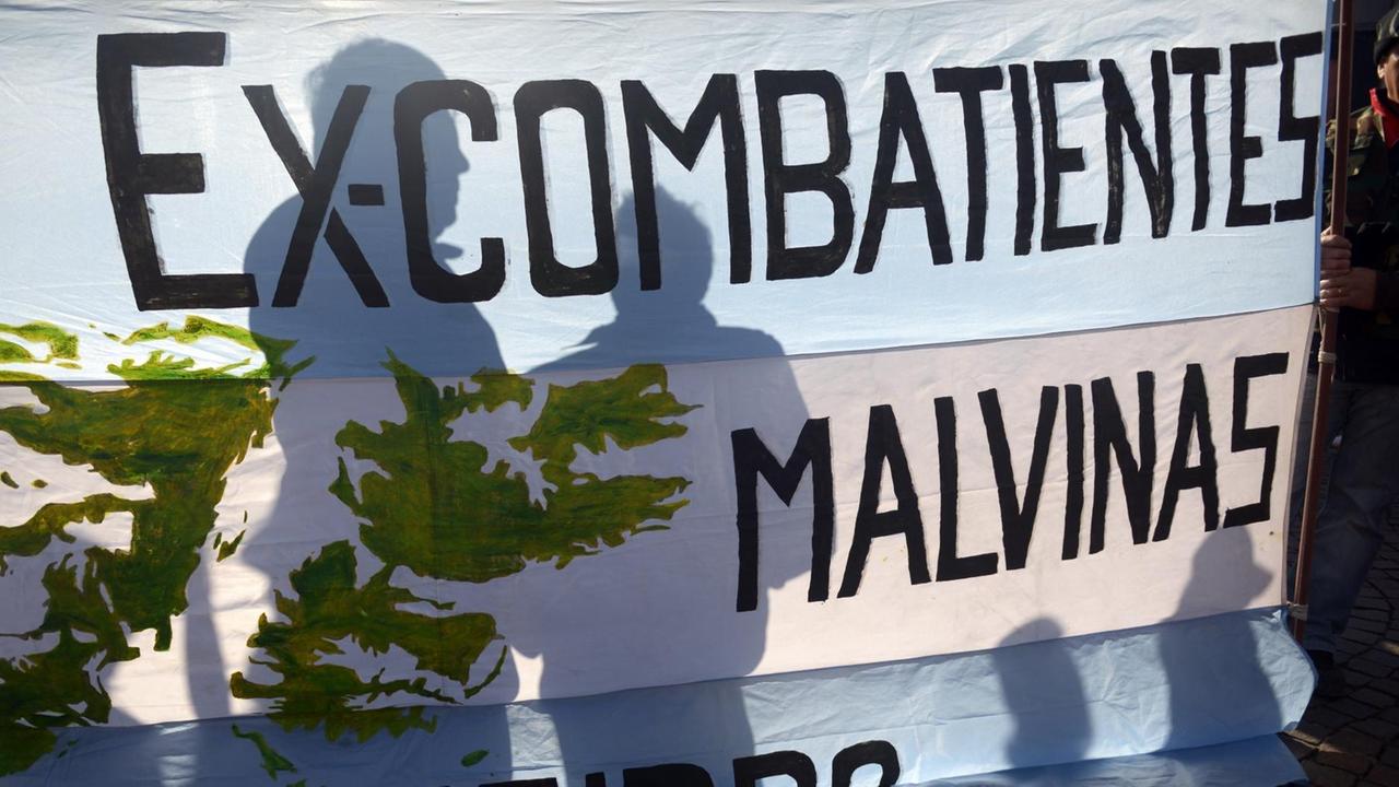 Sie sehen die Schatten von zwei Menschen hinter einer argentinischen Flagge, auf der steht: Ex-Kämpfer Malvinas. Malvinas ist der argentinische Name für die Falkland-Inseln. Sie sehen die Schatten von zwei Menschen hinter einer argentinischen Flagge, auf der steht: Ex-Kämpfer Malvinas. Malvinas ist der argentinische Name für die Falkland-Inseln.