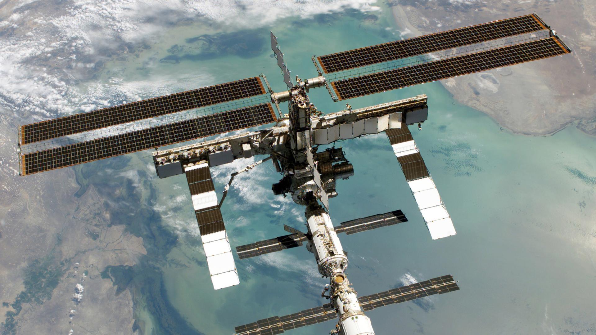 Totale der Internationalen Raumstation, im Hintergrund ein Ausschnitt der Erde, von der Meer und Landflächen zu sehen sind