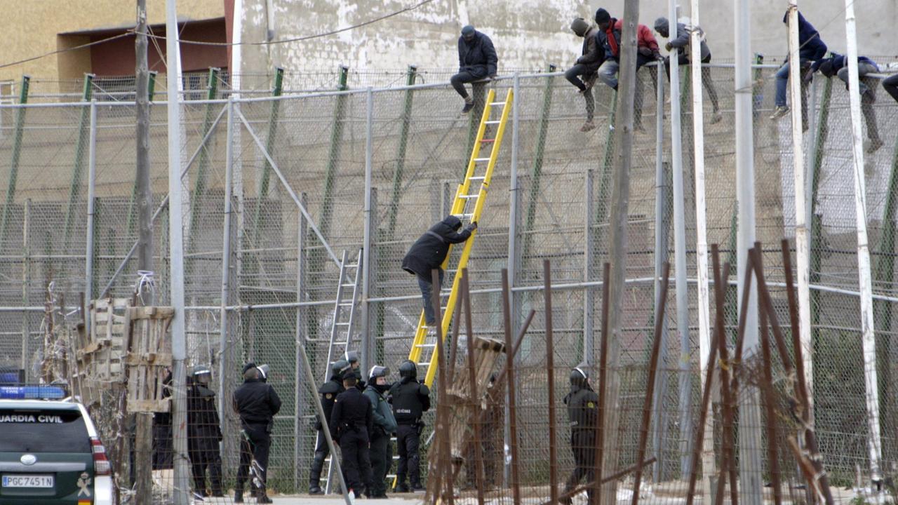 Polizisten beobachten Flüchtlinge, die auf dem Zaun sitzen, der die spanische Exklave Melilla von Marokko trennt.