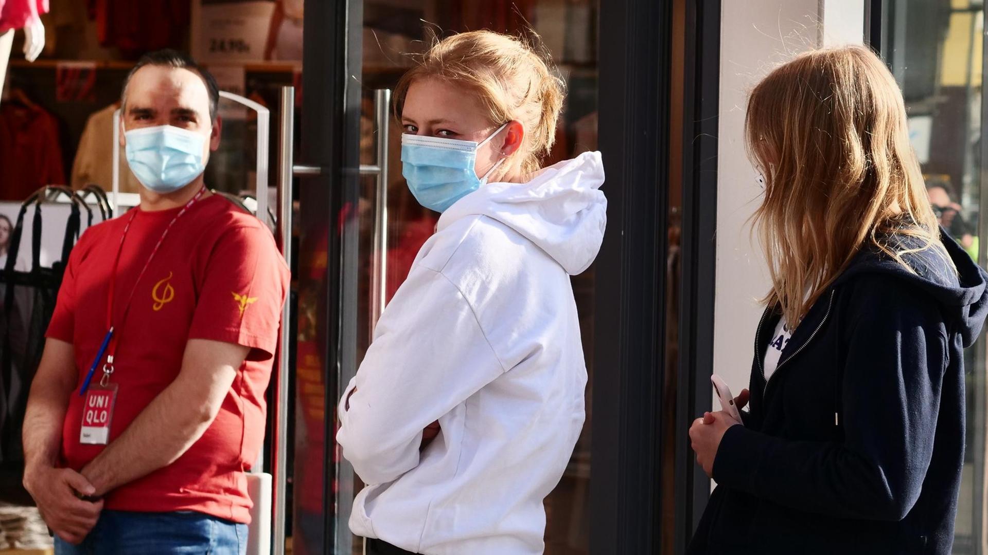 Zwei junge Frauen stehen vor einem Bekleidungsgeschaeft Schlange mit Abstand zueinander und mit Masken gegen das Coronavirus. Zwei junge Frauen stehen vor einem Bekleidungsgeschaeft Schlange mit Abstand zueinander und mit Masken gegen das Coronavirus.