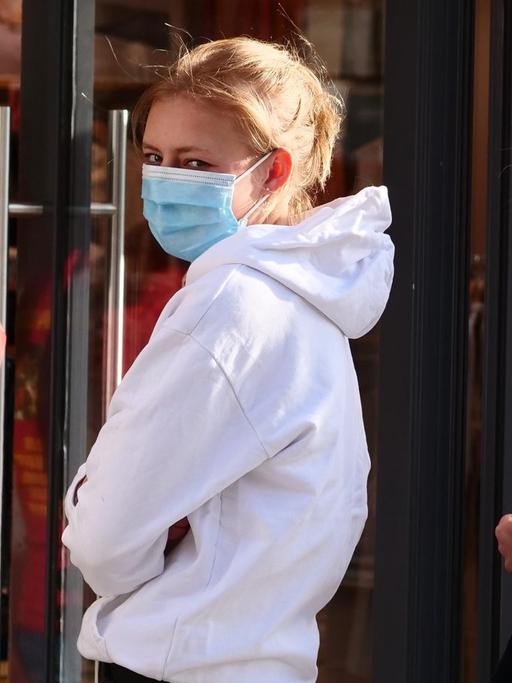 Zwei junge Frauen stehen vor einem Bekleidungsgeschaeft Schlange mit Abstand zueinander und mit Masken gegen das Coronavirus.