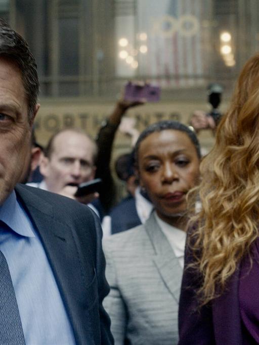 Szene aus der Serie "The Undoing" mit dem Schauspieler Hugh Grant als Jonathan Fraser und der Schauspielerin Nicole Kidman als Grace Fraser. Ein Mann und eine Frau verlassen ein Gerichtsgebäude und werden von der Polizei und Medien begleitet.