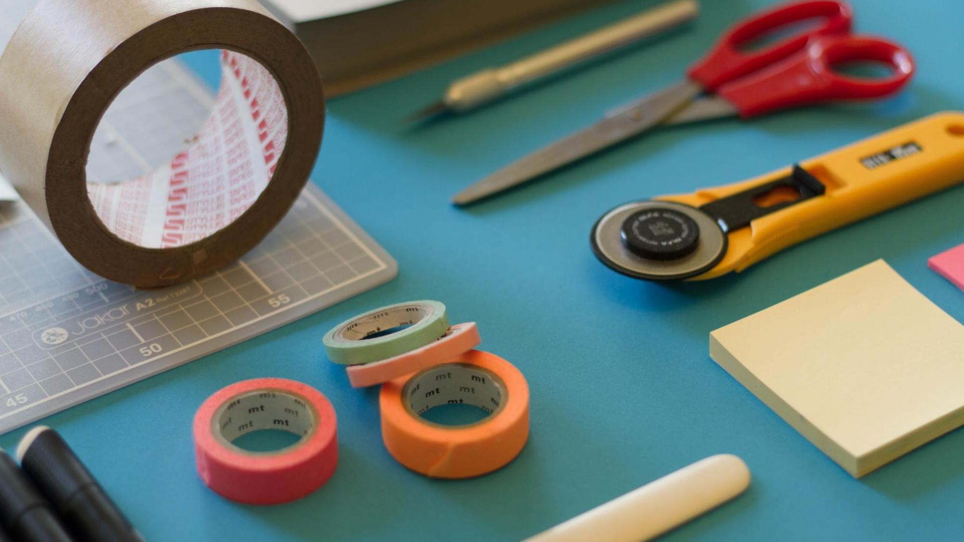 Blick auf eine blaue Tischplatte auf der Schere, Maskingtape, Cutter und weitere Bastelmaterialien angeordnet sind Blick auf eine blaue Tischplatte auf der Schere, Maskingtape, Cutter und weitere Bastelmaterialien angeordnet sind