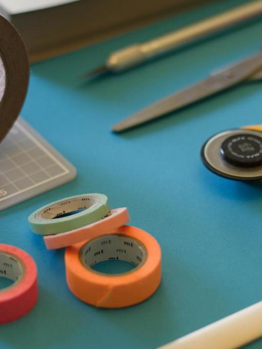 Blick auf eine blaue Tischplatte auf der Schere, Maskingtape, Cutter und weitere Bastelmaterialien angeordnet sind
