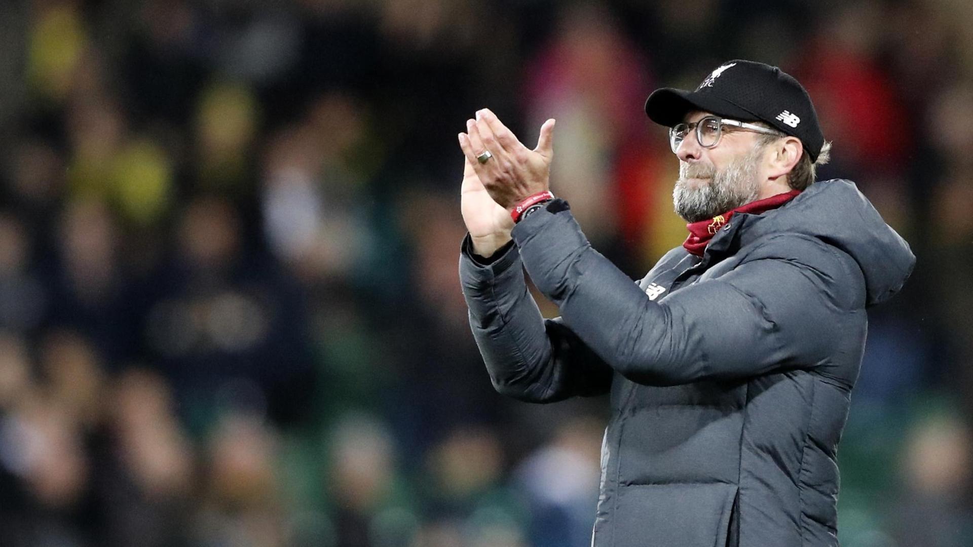 Klopp applaudiert mit zusammengekniffenem Mund. Er trägt eine dunkle Winterjacke, eine schwarze Baseball-Mütze und einen roten Liverpool-Schal. Klopp applaudiert mit zusammengekniffenem Mund. Er trägt eine dunkle Winterjacke, eine schwarze Baseball-Mütze und einen roten Liverpool-Schal.