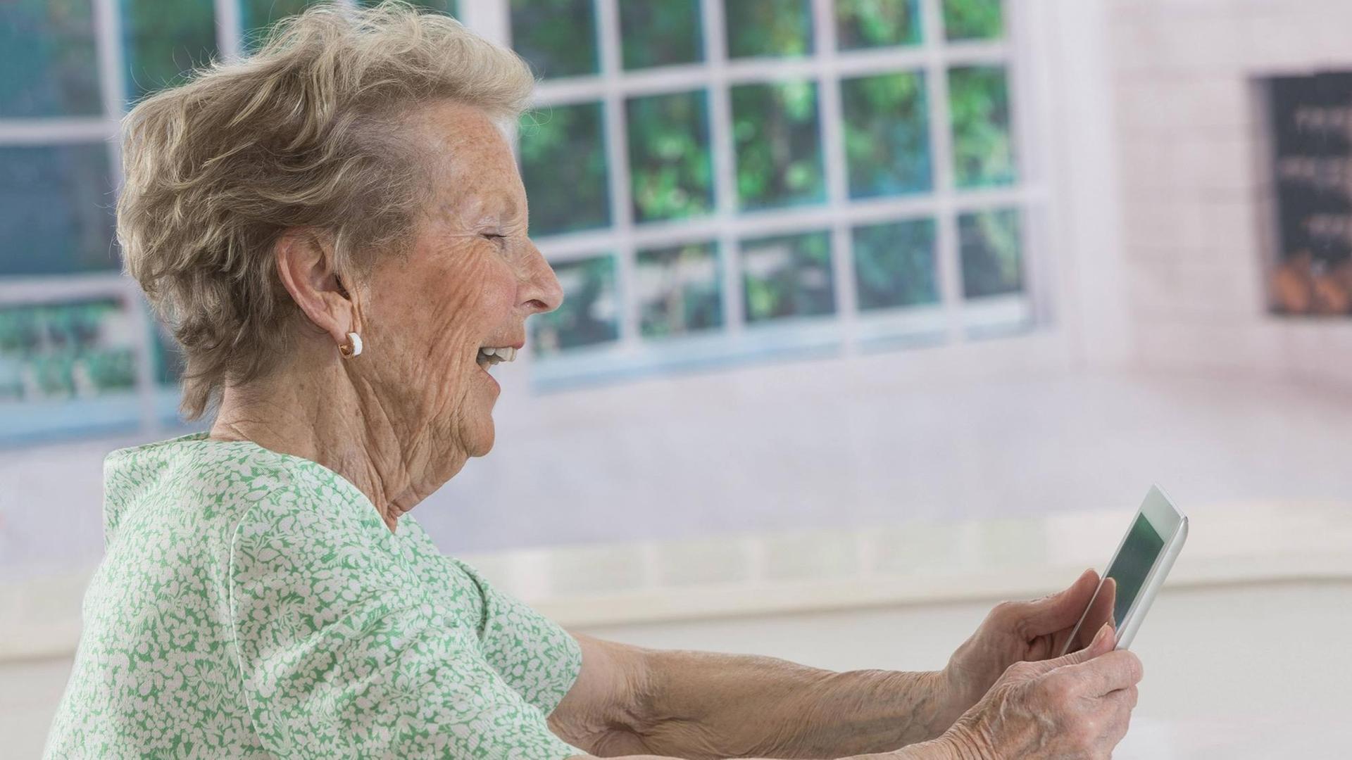 Eine Seniorin sitzt lachend vor einem Tablet, dass sie in beiden Händen hält. 