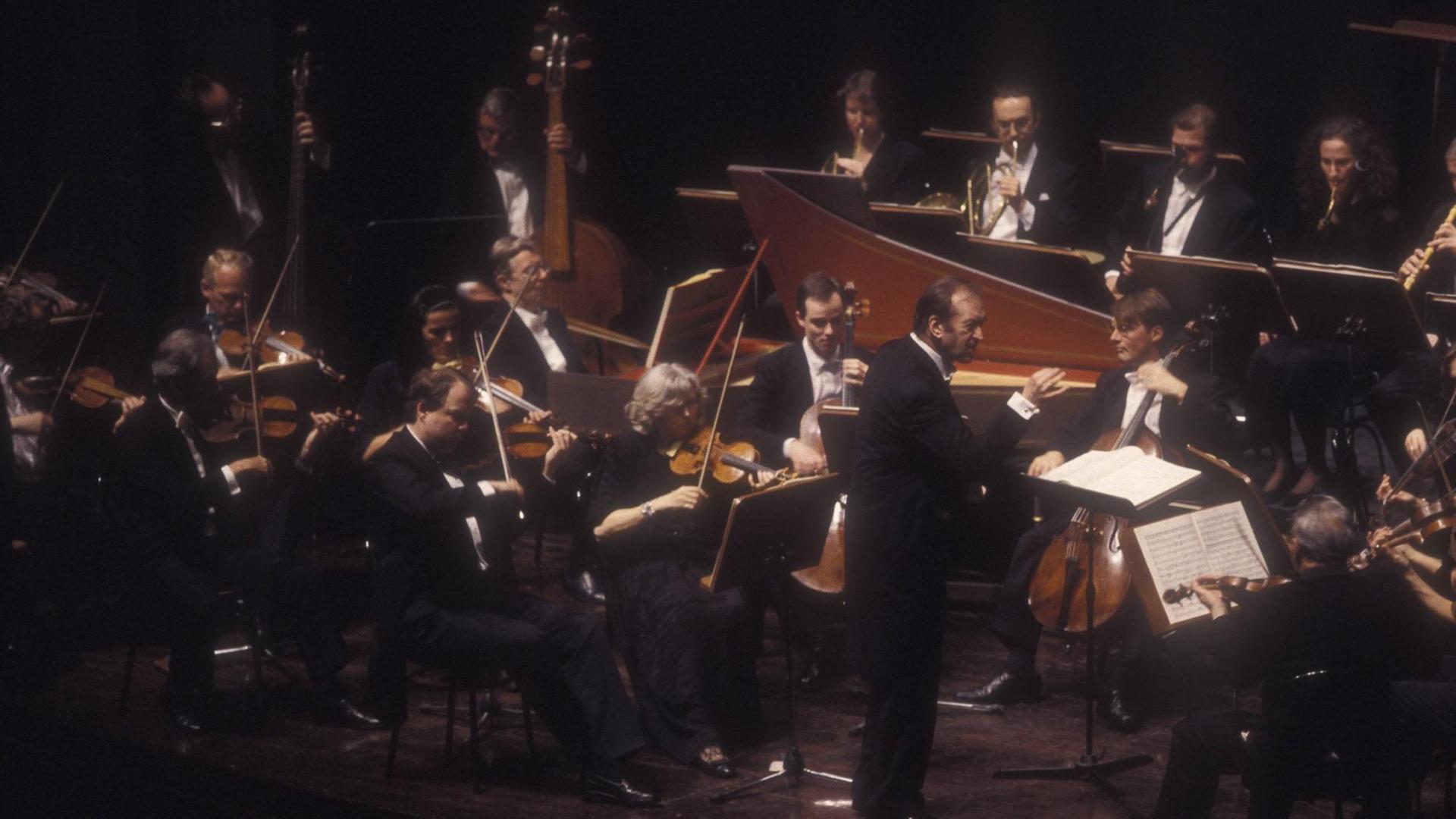 Ein Orchester aus Streichern und Bläsern sitzt in festlicher Kleidung im Halbrund um den Dirigenten, der am Pult steht. Ein Orchester aus Streichern und Bläsern sitzt in festlicher Kleidung im Halbrund um den Dirigenten, der am Pult steht.