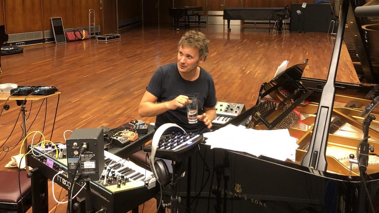 Ein junger Mann sitzt vor einem Flügel, rechts und links davon Synthesizer. Er hält ein Wasserglas in der Hand. Ein junger Mann sitzt vor einem Flügel, rechts und links davon Synthesizer. Er hält ein Wasserglas in der Hand.