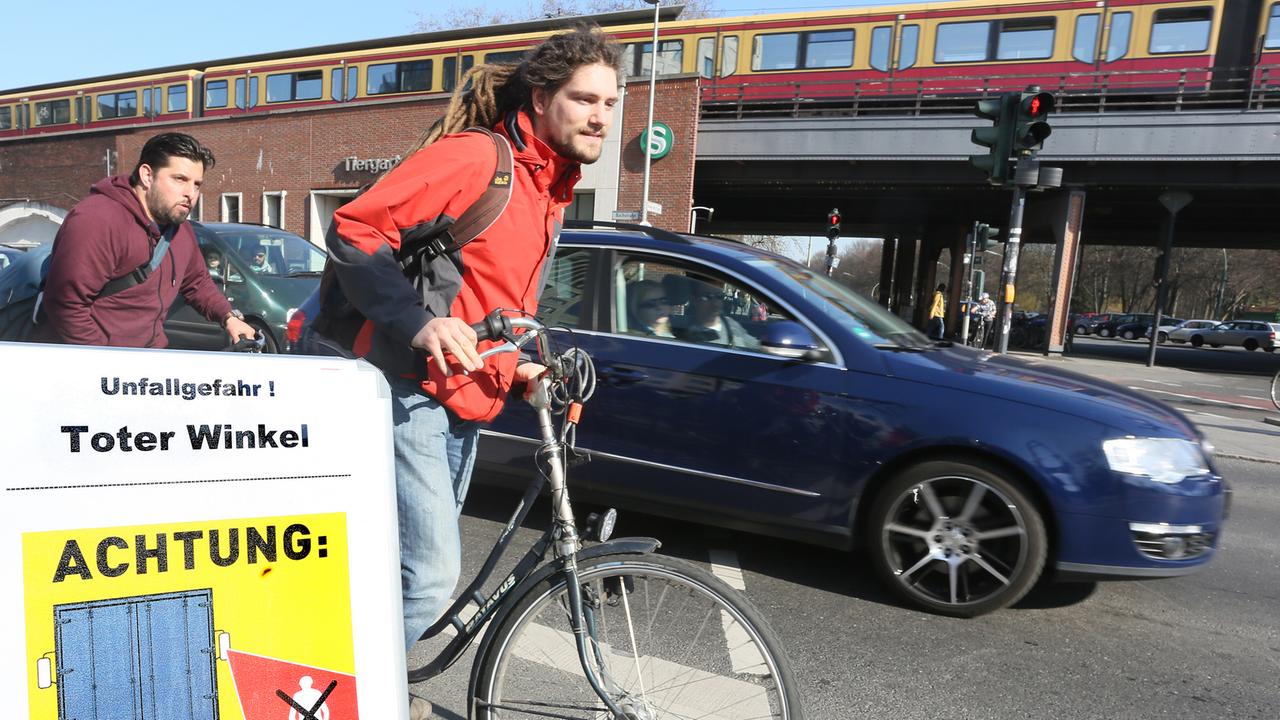 Fahrradfahrer fahren auf einem Fahrradweg an einer Kreuzung in Berlin geradeaus, während ein Auto rechts abbiegen will, davor weist ein Schild über den Toten Winkel als Unfallgefahr hin. Fahrradfahrer fahren auf einem Fahrradweg an einer Kreuzung in Berlin geradeaus, während ein Auto rechts abbiegen will, davor weist ein Schild über den Toten Winkel als Unfallgefahr hin.