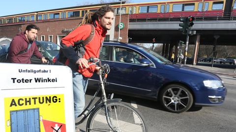 Fahrradfahrer fahren auf einem Fahrradweg an einer Kreuzung in Berlin geradeaus, während ein Auto rechts abbiegen will, davor weist ein Schild über den Toten Winkel als Unfallgefahr hin.