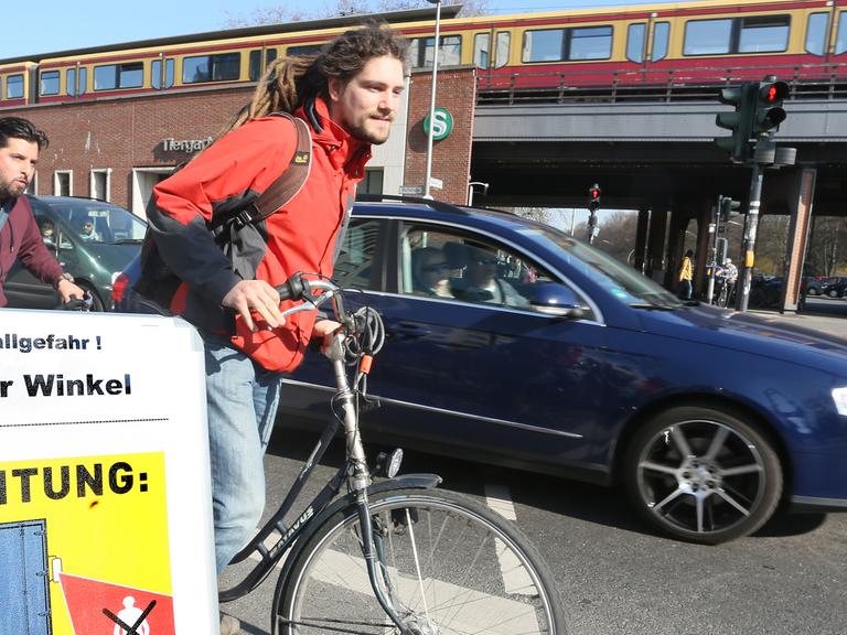 Fahrradfahrer fahren auf einem Fahrradweg an einer Kreuzung in Berlin geradeaus, während ein Auto rechts abbiegen will, davor weist ein Schild über den Toten Winkel als Unfallgefahr hin.