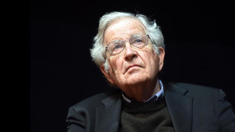 Noam Chomsky schaut vor schwarzem Hintergrund während einer Rede nach oben.