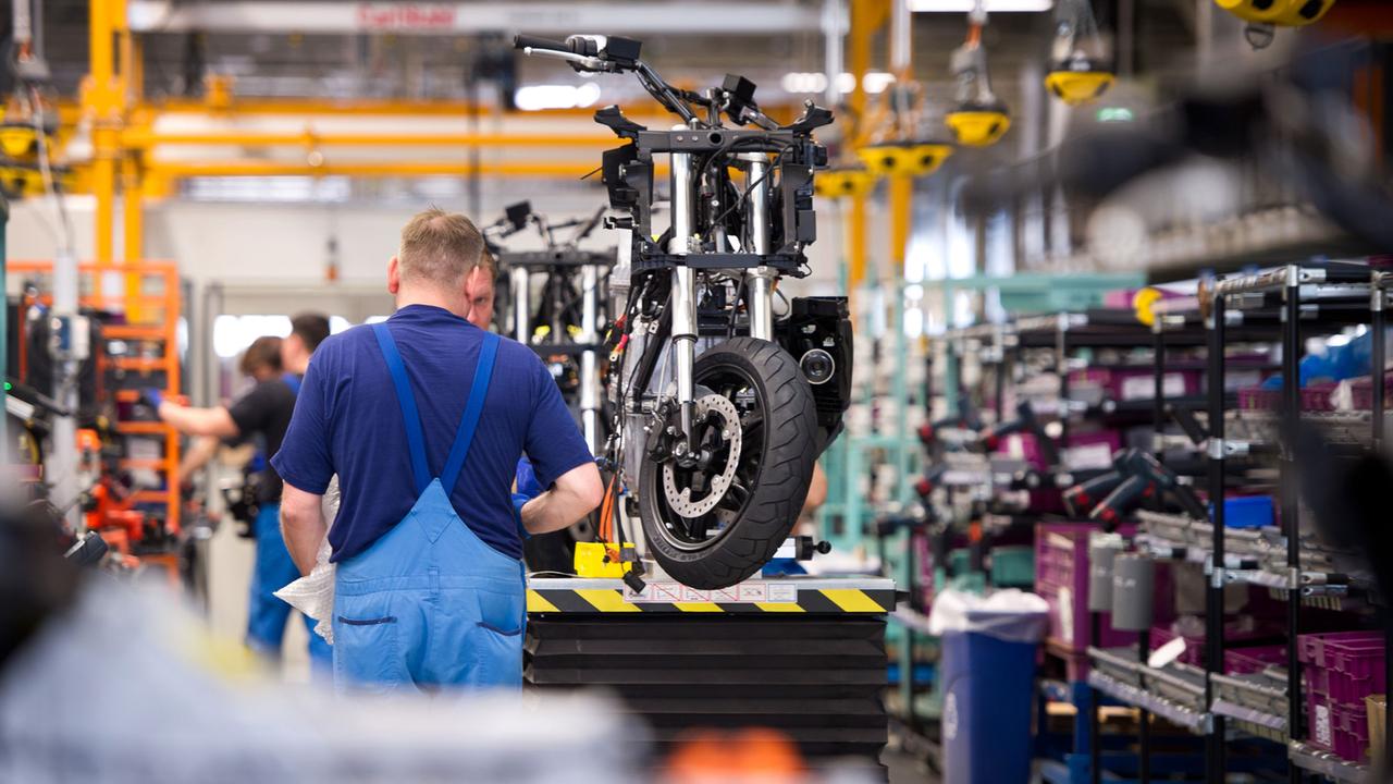 Ein Monteur steht im Berliner BMW-Werk in einer Halle und spricht mit einem Kollegen. Neben ihm ist ein Elektroroller zu sehen, der gerade montiert wird. Ein Monteur steht im Berliner BMW-Werk in einer Halle und spricht mit einem Kollegen. Neben ihm ist ein Elektroroller zu sehen, der gerade montiert wird.