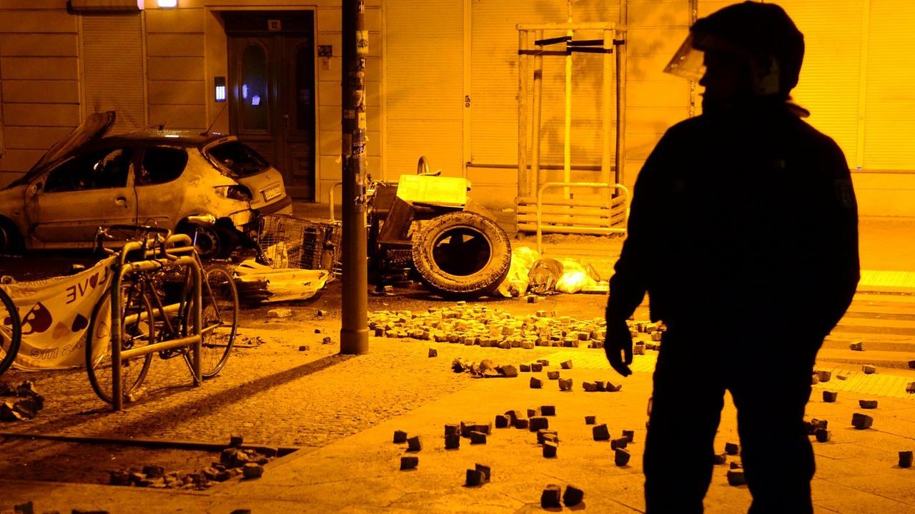 Die Silhouette eines Polizisten in Kampfmonitur in der gelblich beleuchteten Straße. Im Hintergrund ein ausgebranntes Auto und viele Pflasterssteine. Die Silhouette eines Polizisten in Kampfmonitur in der gelblich beleuchteten Straße. Im Hintergrund ein ausgebranntes Auto und viele Pflasterssteine.