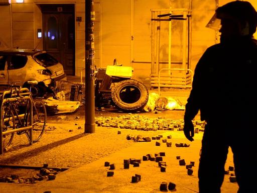 Die Silhouette eines Polizisten in Kampfmonitur in der gelblich beleuchteten Straße. Im Hintergrund ein ausgebranntes Auto und viele Pflasterssteine.