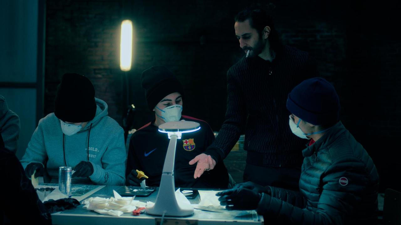 Die Filmszene aus der Serie "Mocro Maffia" zeigt eine Drogenlabor. In einem spärlich beleuchteten Raum sitzen jungen Männer mit Gesichtsmasken, ein älterer Mann gibt ihnen Anweisungen. Die Filmszene aus der Serie "Mocro Maffia" zeigt eine Drogenlabor. In einem spärlich beleuchteten Raum sitzen jungen Männer mit Gesichtsmasken, ein älterer Mann gibt ihnen Anweisungen.