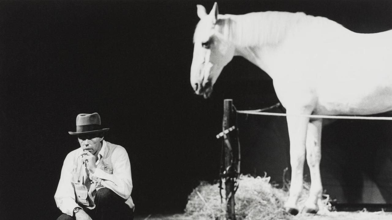 Joseph Beuys kniet und hinter ihm steht ein weißes Pferd