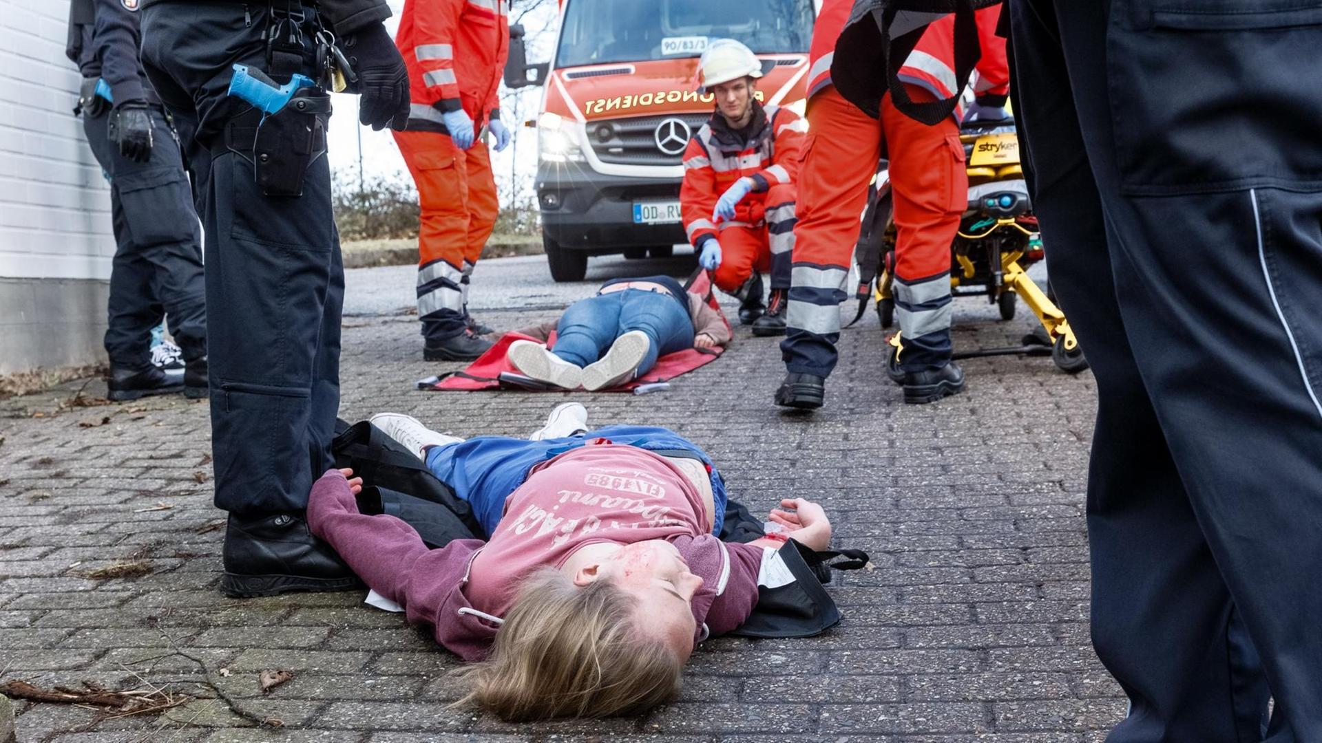 Ein Kind und eine erwachsene Person liegen offenbar verletzt auf dem Asphalt. Zu sehen sind drumherum die Beine von Polizisten und Feuerwehrmännern in ihren Uniformen. Ein Feuerwehrmann ist in die Hocke gegangen. Ein Kind und eine erwachsene Person liegen offenbar verletzt auf dem Asphalt. Zu sehen sind drumherum die Beine von Polizisten und Feuerwehrmännern in ihren Uniformen. Ein Feuerwehrmann ist in die Hocke gegangen.